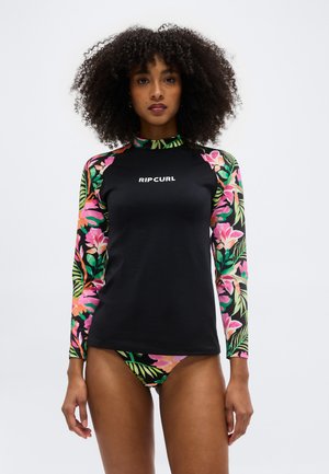 Kobieta o kręconych włosach, ubrana w czarną koszulkę do pływania z rękawami w kwiatowy wzór oraz pasujące kwiatowe bikini, na jednolitym tle.