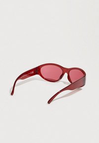 Κόκκινα γυαλιά ηλίου με οβάλ φακούς. Κατασκευασμένα από γυαλιστερό πλαστικό, με λεπτούς βραχίονες και διακριτικό λογότυπο στο σκελετό. Ελαφρώς κυρτό σχήμα.