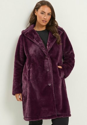 Femme portant un long manteau en velours violet foncé boutonné avec un large col, debout devant un fond clair uni.