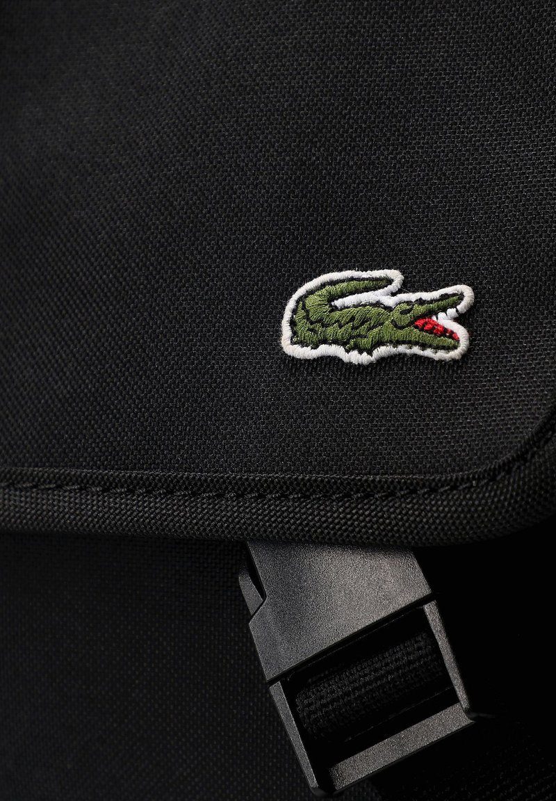 Sac d'épaule en tissu noir avec un petit logo crocodile vert et blanc, équipé d'une boucle en plastique noir et d'une surface texturée.