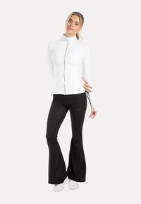 White zip-up athletic jacket with high collar, paired with black flared yoga pants. Fabric appears smooth with fitted design. White sneakers visible.