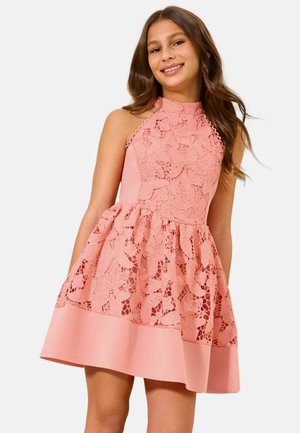 Junge Frau lächelt, trägt ein ärmelloses rosa Spitzenkleid mit floralem Muster und ausgestelltem Rock, steht vor einem schlichten Hintergrund.