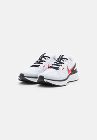 Bijele atletske tenisice s mrežastim gornjim dijelom, crnim vezicama i crvenim Nike odznačenjem. Imaju crno-bijelu potplatu s teksturiranim uzorkom.