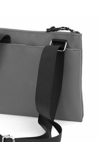 Šedá textilní taška s hladkou texturou, vybavená černým nastavitelným popruhem a stříbrnými kovovými detaily. Horní uzavírání na zip a boční kapsa.