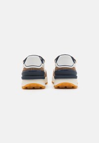 Zapatillas con parte superior de gamuza marrón y malla azul marino, acentos blancos y una suela de goma gruesa en crema claro y naranja. La vista trasera muestra un collar acolchado.