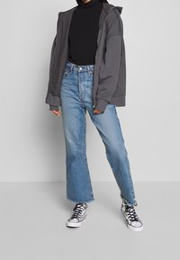 Sudadera gris con cremallera, cuello de tortuga negro, vaqueros de pierna ancha de cintura alta en azul claro, y zapatillas de deporte con patrón en blanco y negro. Conjunto casual.