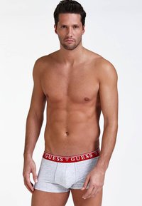 Boxeri gri cu un bandă roșie cu logo-ul "GUESS", ajustați, dintr-un material neted. Modelul prezentat stă fără tricou, pe un fundal neutru.