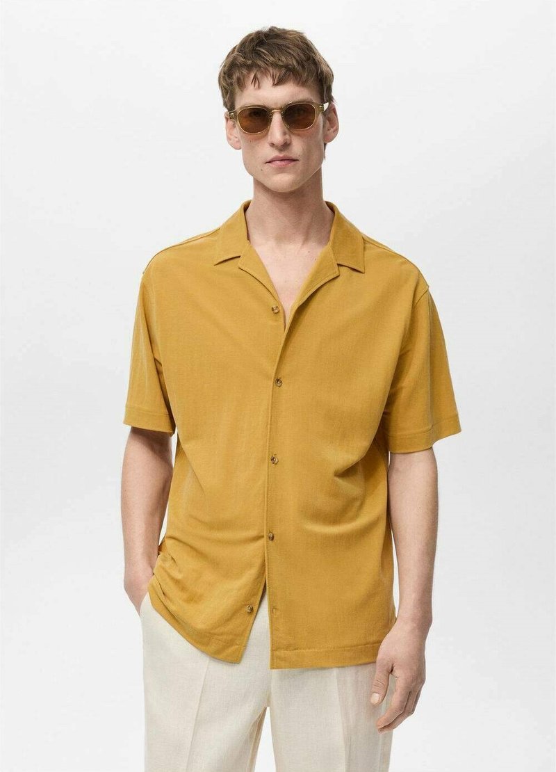 Mustard yellow short-sleeve shirt with a relaxed fit, small collar, and button closure, paired with light beige pants and sunglasses.