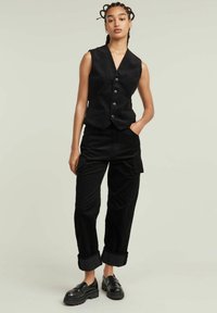 Zwarte corduroy gilet met een V-hals, knoopsluiting en voorzakken, gecombineerd met zwarte cargobroek met opgerolde boorden en zijzakken.