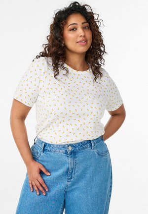 Frau mit lockigem Haar, die ein weißes T-Shirt mit Zitronenmuster und hoch taillierte blaue Jeans trägt, steht mit den Händen in der Hüfte.