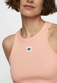 Vrouw in een mouwloze perzikkleurige ribben tanktop met een klein vierkant patch op de borst met daarop twee horizontale zwarte lijnen.