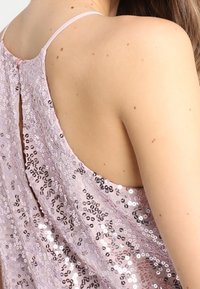 Haut rose clair à sequins avec un dos dégagé. Doté de fines bretelles et d'une fermeture à boutons dans le haut du dos. Surface texturée et étincelante.