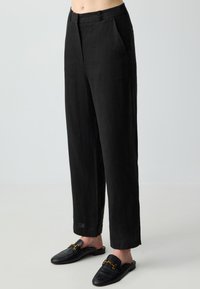Schwarze Leinenhose mit geradem Schnitt, ausgestattet mit zwei Seitentaschen und einem schlichten Bund. Kombiniert mit flachen schwarzen Slippern.