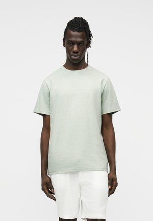 Uomo dalla pelle scura con capelli intrecciati che indossa una maglietta a maniche corte grigio chiaro e pantaloncini bianchi, in piedi davanti a uno sfondo semplice.