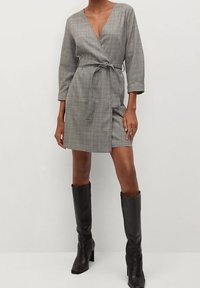 Szara w kratę sukienka o kroju wrap z głębokim dekoltem w literę V, długimi rękawami i wiązaniem w pasie, zestawiona z czarnymi botkami do kolan. Gładka tekstura materiału.
