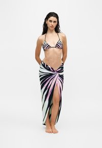 Femme debout pieds nus, portant un haut de bikini à rayures zèbre et une jupe portefeuille assortie fendue devant, sur fond blanc uni.