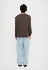 Maglietta lunga marrone a maniche lunghe con collo rotondo, abbinata a jeans larghi blu chiaro. La maglietta presenta un sottile dettaglio testurizzato sul retro.