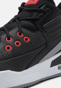 Gros plan d’une basket noire montrant la texture du cuir, des lacets noirs passés dans des œillets rouges, et une partie de la semelle blanche et grise.
