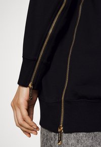 Felpa nera con dettagli della cerniera dorati sui polsini, orlo a coste e una texture di tessuto liscia. I cursori della cerniera presentano il marchio.