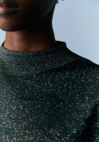 Top verde scuro scintillante con colletto alto, caratterizzato da una superficie texturizzata di fili luccicanti e una silhouette aderente.