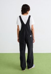 Schwarze Denim-Latzhose mit weitem Bein und verstellbaren Trägern, ausgestattet mit zwei hinteren Taschen und einem kleinen Markenlabel auf der rechten Tasche.