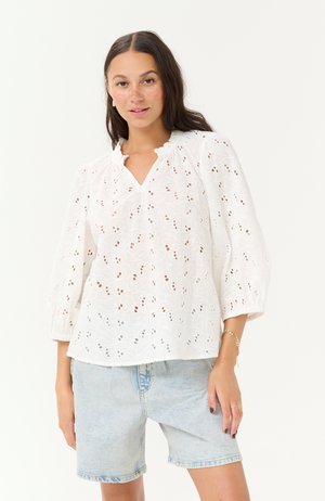 Weiße Bluse mit floralem Stickereimuster, V-Ausschnitt und sanften Puffärmeln. Getragen mit hellen Jeansshorts.