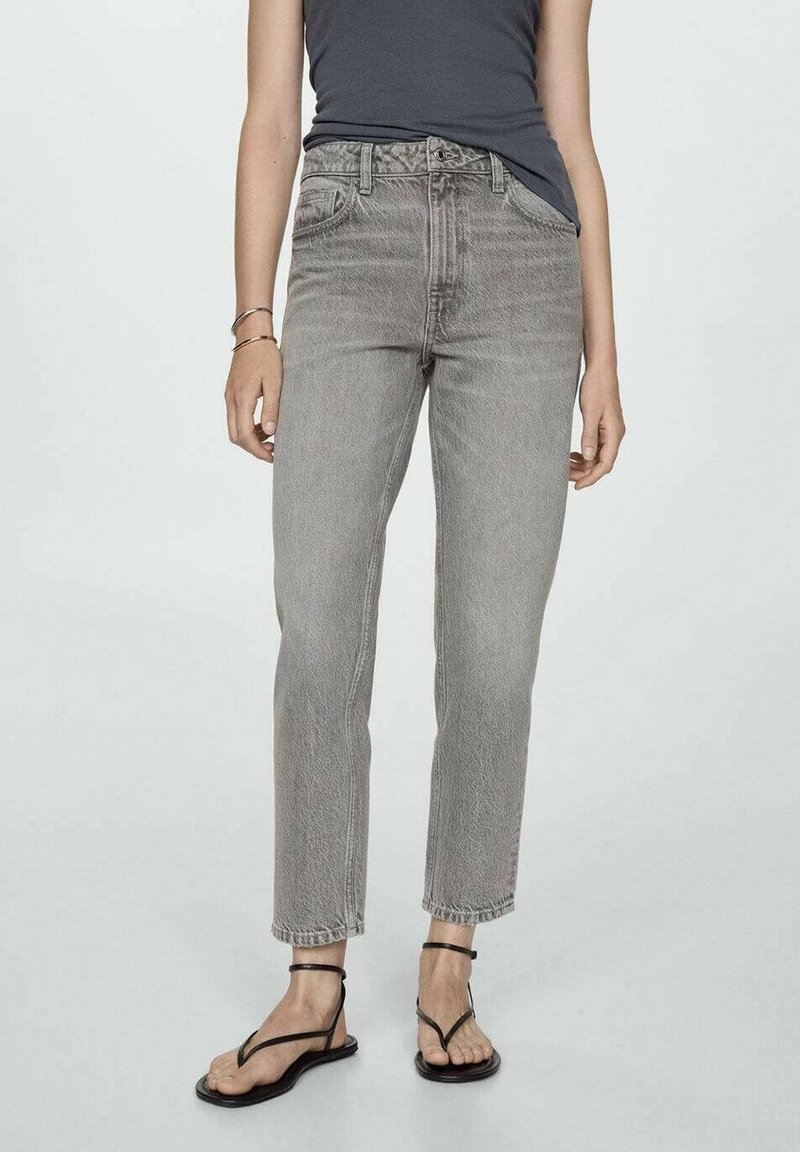 Personne portant un jean gris délavé taille haute et des sandales noires à brides, debout devant un fond clair et uni.
