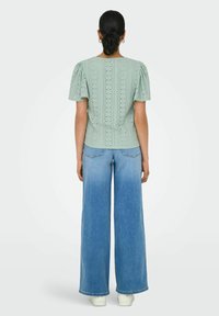 Helles grünes Oberteil mit Puffärmel, Lochmusterung und abgerundetem Saum, kombiniert mit weit geschnittenen blauen Jeans und weißen Sneakers.