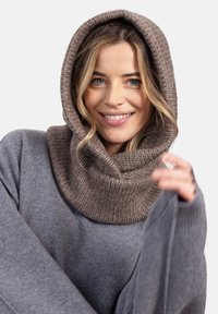 Uśmiechnięta kobieta z luźnymi blond włosami nosi taupe'owy dziany kaptur oraz szary sweter z długim rękawem, trzymając jeden rękaw blisko swojej twarzy.