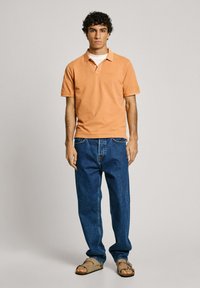 Oranžs kokvilnas polo krekls ar apkakli, apvienots ar brīviem ziliem džinsiem un bēšām sandālēm. Modelis stāv uz neitrāla fona.