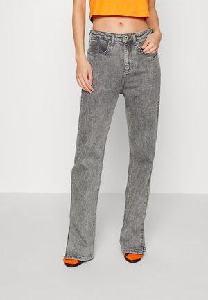 Vrouw draagt hoge taille, grijze acid-wash wijde jeans, cropped oranje top en zwarte sandalen met feloranje sokken, staand tegen een witte achtergrond.