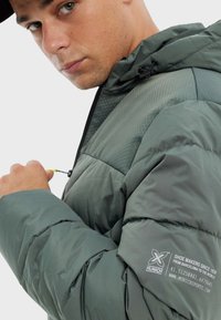 Veste doudoune verte avec un motif en nid d'abeille texturé sur la partie supérieure, dotée d'une fermeture éclair et de détails de logo sur la manche.