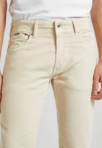 Pantalon en velours côtelé beige avec une coupe slim, doté de poches avant, passants pour ceinture et d'une texture côtelée subtile. Fermeture par bouton et fermeture éclair.
