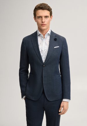 Man met kort krullend haar draagt een marineblauw colbert en broek over een wit overhemd met patroon, staand tegen een effen lichte achtergrond.