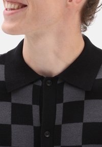 Pánský polo shirt s černobílým kostkovaným vzorem, plochým límcem, pěti knoflíky a jemnou texturou. Zaměření na horní část a výstřih.
