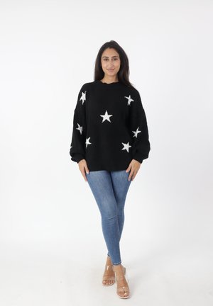 Zwarte gebreide trui met witte sterrenpatronen, oversized pasvorm, geribbelde manchetten, gecombineerd met blauwe skinny jeans en doorzichtige haksandalen.