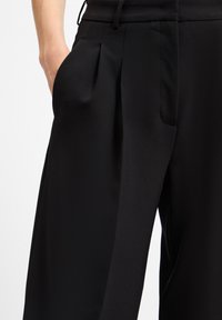 Zwarte broeken met een gladde textuur, plooien, zijzakken en een getailleerde pasvorm, die een strakke en moderne uitstraling benadrukken.