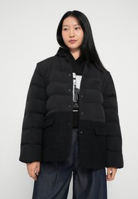 Melna pūkainā jaka ar teksturētu melnu apakšējo paneli, spiedpogu aizdari un sānu kabatām. Valkāta virs melna kapuces džempera un džinsu biksēm.