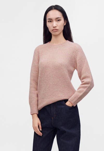 Jeune femme aux longs cheveux noirs portant un pull en laine rose clair et un jean bleu foncé, debout avec une main dans la poche.