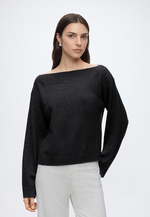 Femme aux cheveux foncés portant un pull noir à épaules dénudées et un pantalon blanc, debout devant un fond clair uni.