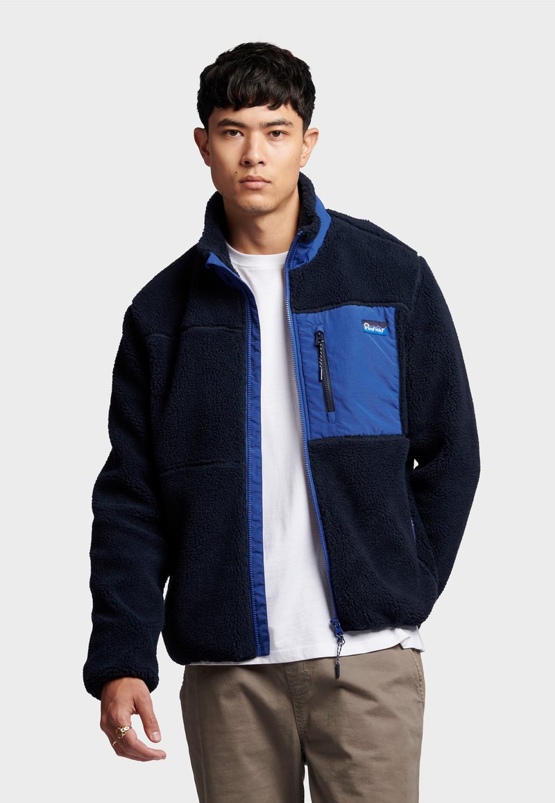 Fleecejacke in Marineblau mit einem blauen Einsatz, durchgehendem Reißverschluss, Reißverschlusstasche auf der Brust und strukturiertem Stoff. Mit Stehkragen und lockerer Passform.