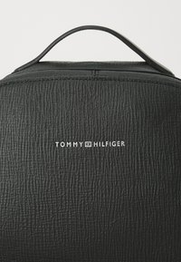 Must mustriga seljakott, millel on ülemine käepide ja valge "TOMMY HILFIGER" logo. Disain rõhutab minimalistlikku stiili peente mustritega.
