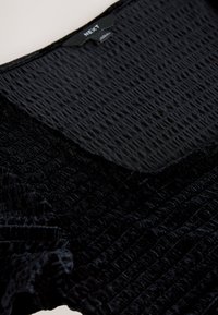 Sort tekstureret top med krøllet stof og en glat sektion ved halsudskæringen, med synligt mærke i midten.