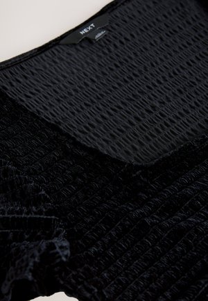 Czarny teksturowany top wykonany z marszczonej tkaniny z gładkim fragmentem przy dekolcie, z widoczną oznaką marki na środku.