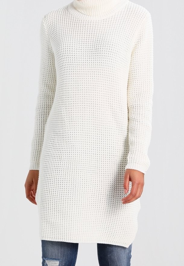 Weißes, gestricktes Sweatkleid mit hohem Kragen, das ein lockeres, strukturiertes Design, lange Ärmel und eine Oberschenkellänge aufweist. Kombiniert mit verwaschenen Jeans.