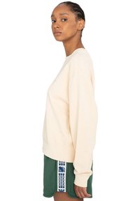Beiger Sweatshirt mit Rundhalsausschnitt, gerippten Bündchen und Saum. Kombiniert mit grünen Shorts, die ein blaues gemustertes Seitenpanel haben.