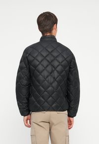 Giubbotto invernale nero con motivo a rombi, dotato di colletto alto e polsini elasticizzati, indossato sopra pantaloni kaki.