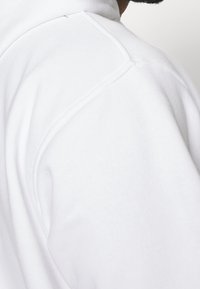 Felpa bianca in tessuto morbido e testurizzato. Presenta cuciture piatte e un colletto alto, evidenziando un design pulito e minimalista.