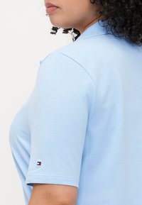 Hele sinine polosärk, mis on valmistatud struktureeritud kangast, lühikeste varrukatega ja millel on väike logoaplikatsioon varrukal punases, valges ja sinises värvitoonis.