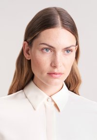 Beige collared shirt with a smooth, soft texture. Features a button-down front and a classic, tailored design with a structured collar.